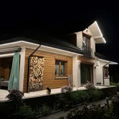 Nocne oświetlenie podkreśla nierówności na elewacji pod światło, tworząc ciekawy efekt na drewnianej części domu i stosie drewna.