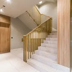 Nowoczesna klatka schodowa z białym, kamiennym tynkiem mozaikowym, złotymi balustradami i drewnianymi elementami. Obok winda.