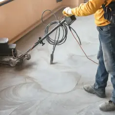 Pracownik wykonuje szlifowanie posadzki betonowej za pomocą maszyny. Widać kurz i ślady po obróbce.