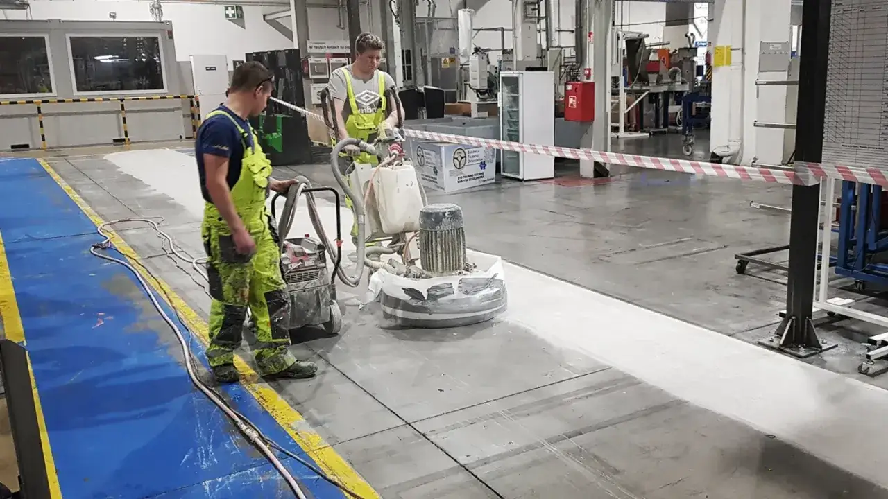 Dwóch pracowników w zielonych ogrodniczkach wykonuje szlifowanie posadzki betonowej za pomocą maszyn.