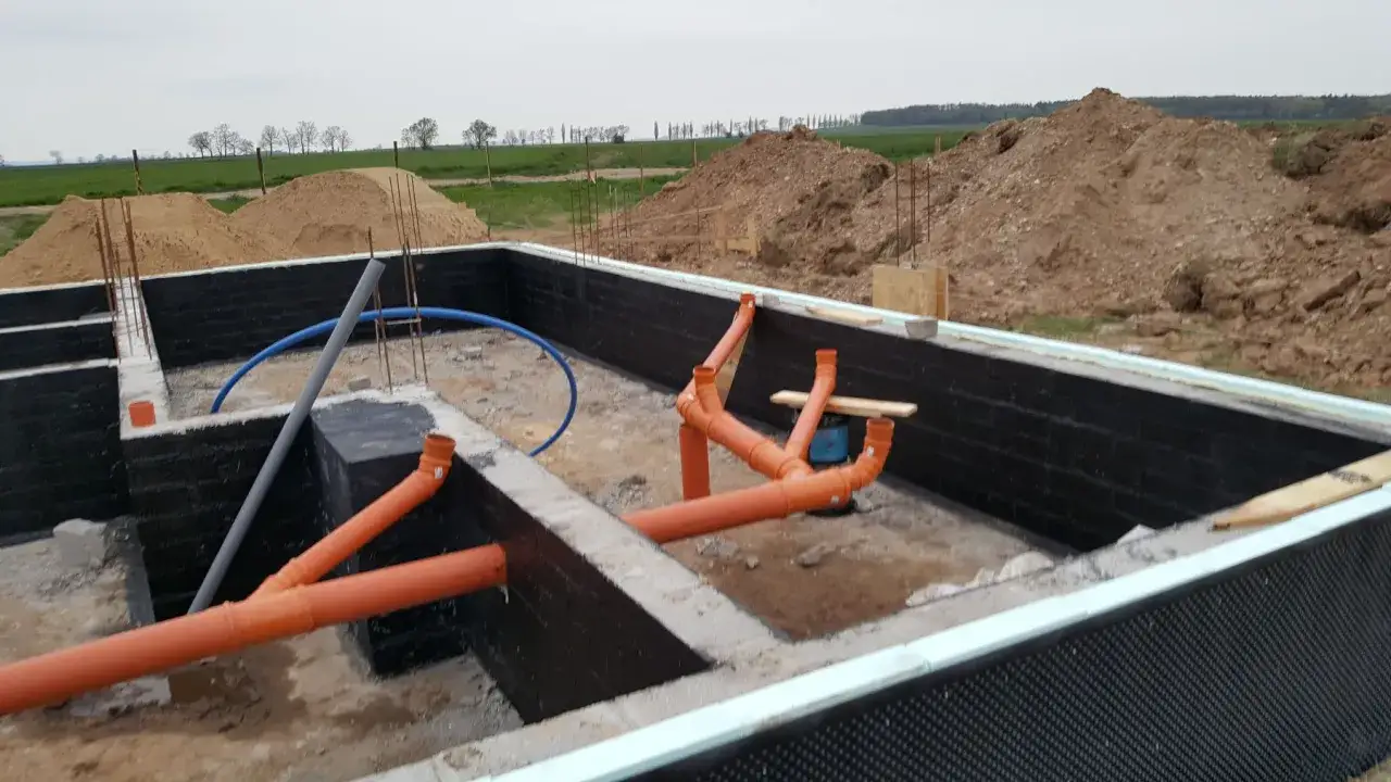 Pomarańczowe rury tworzą skomplikowane przyłącze kanalizacyjne w wykopie fundamentowym.