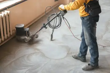 Pracownik wykonuje szlifowanie posadzki betonowej za pomocą maszyny. Widać kurz i ślady po obróbce.