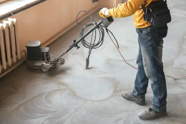 Pracownik wykonuje szlifowanie posadzki betonowej za pomocą maszyny. Widać kurz i ślady po obróbce.